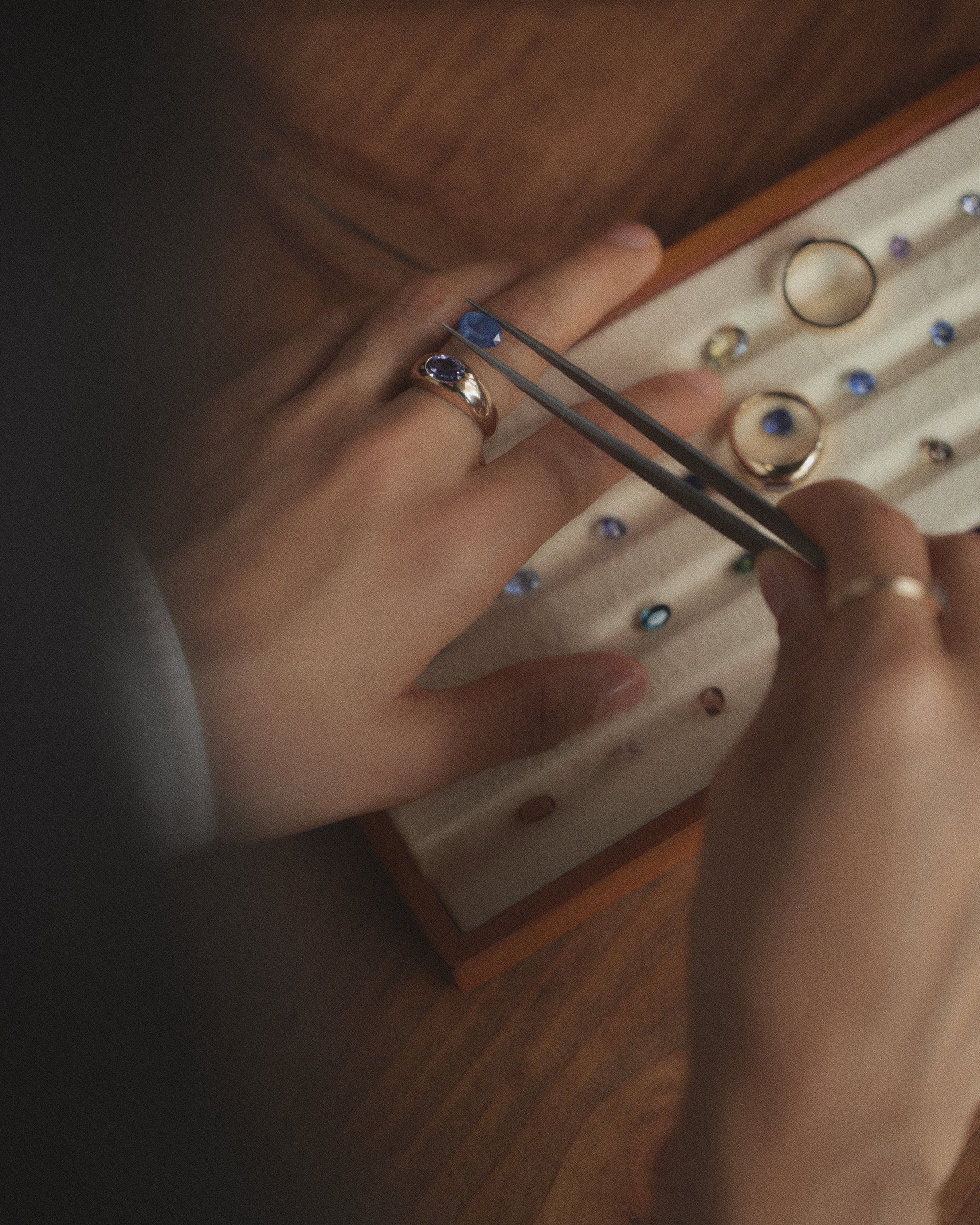 [lifestyle]Close-up of hands with rings using tweezers over a wooden jewelry box with rings.